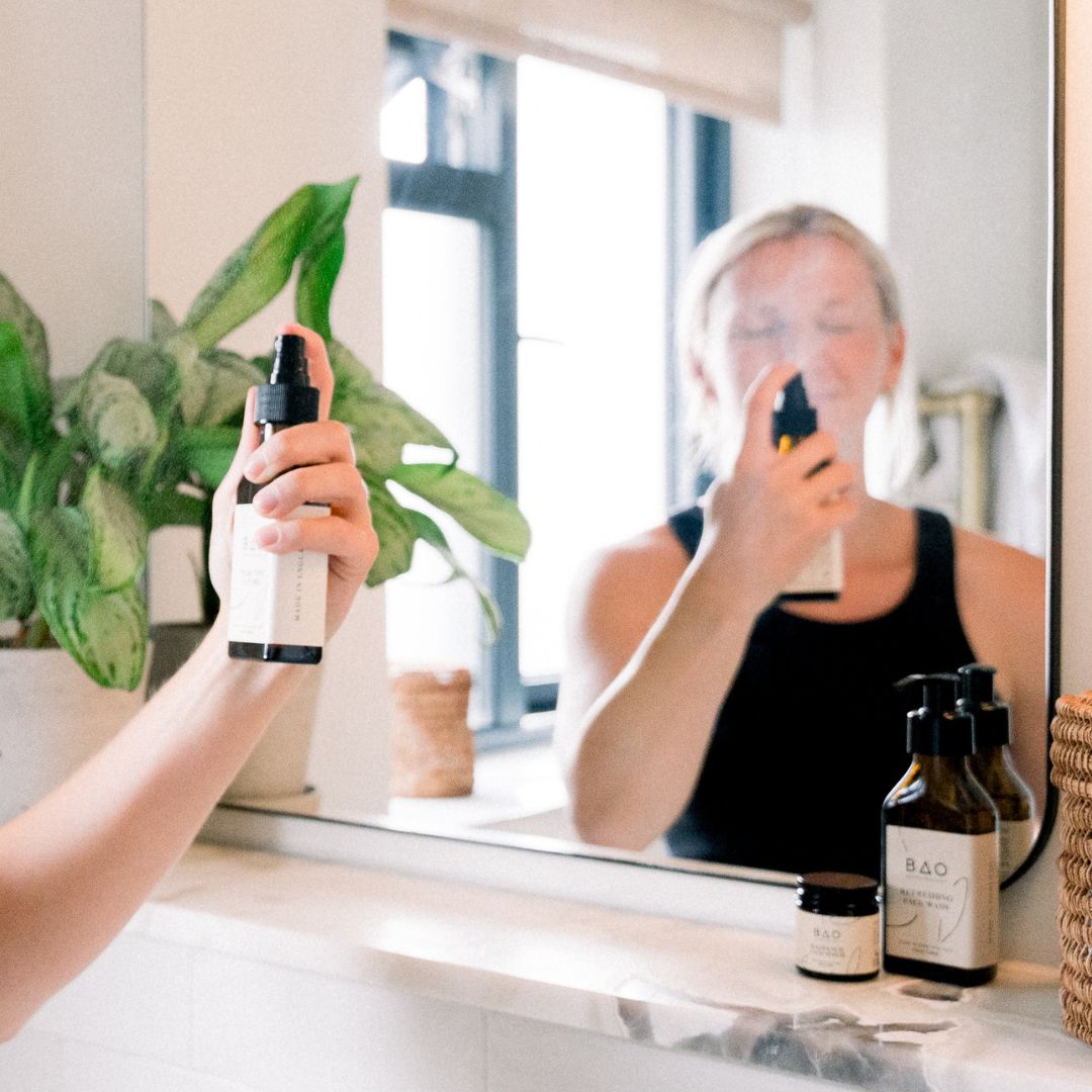 Woman spraying BAO Hydrating Face Mist onto her face in front of a bathroom mirror