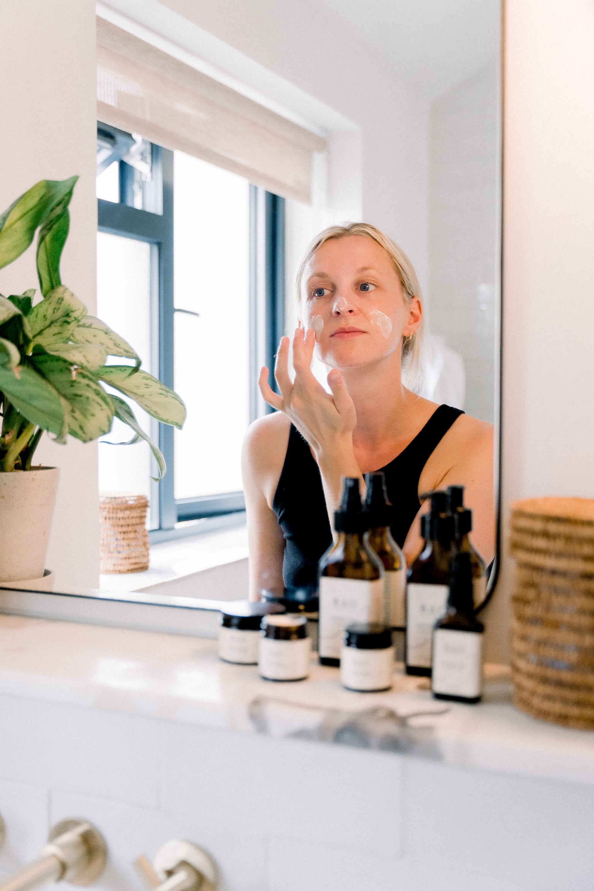 Woman applying skincare product in front of a mirror with various bottles on the counter.