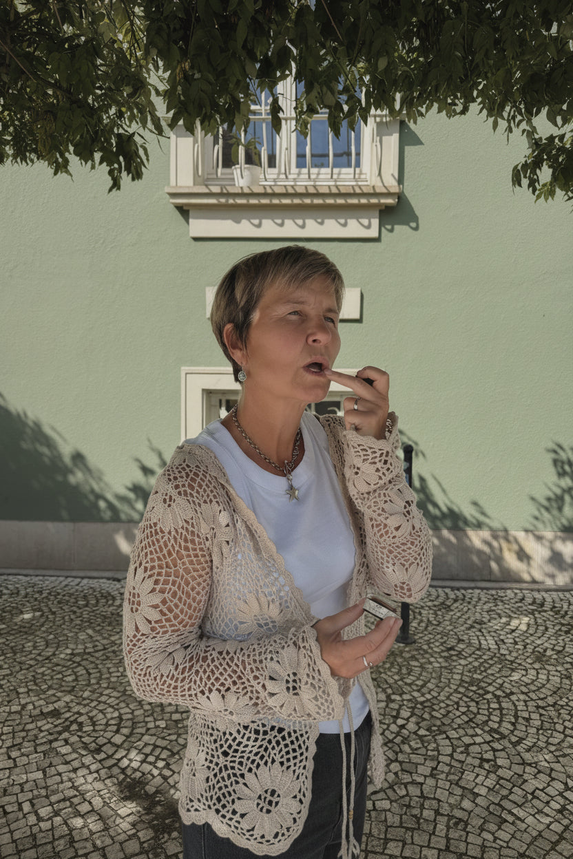 A woman with short blonde hair stands outdoors in front of a light green building, wearing a white t-shirt, a cream-colored crochet cardigan, and a gold necklace while holding a small white item.