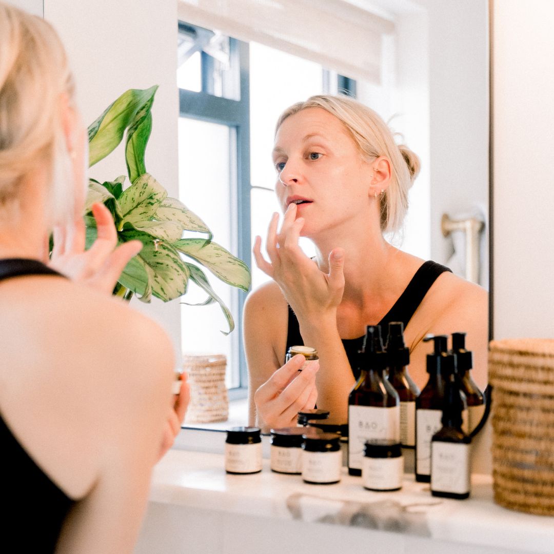 Woman applying BAO Skin Saviour Balm to her lips in front of a bathroom mirror