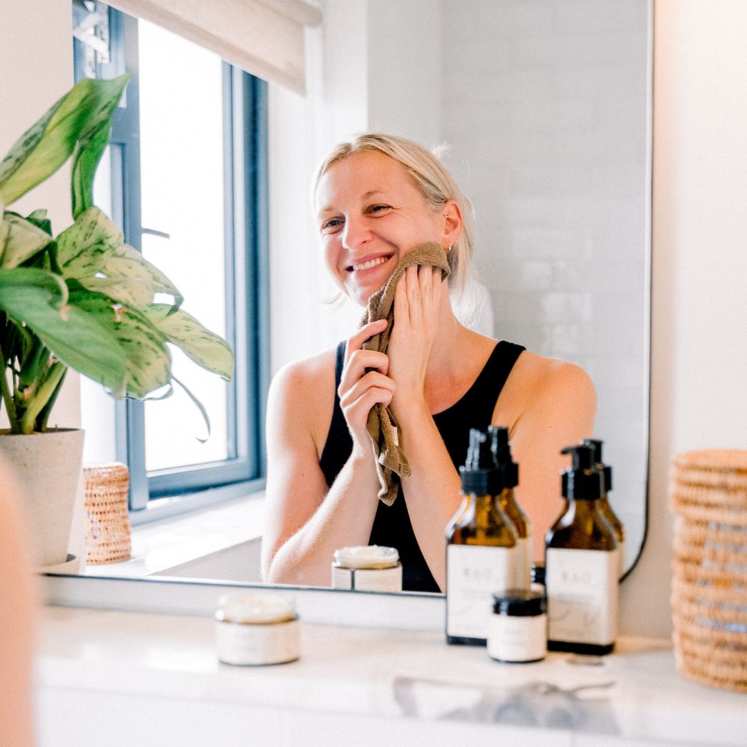 Woman smiling while wiping her face with a towel at a bathroom mirror with BAO products nearby