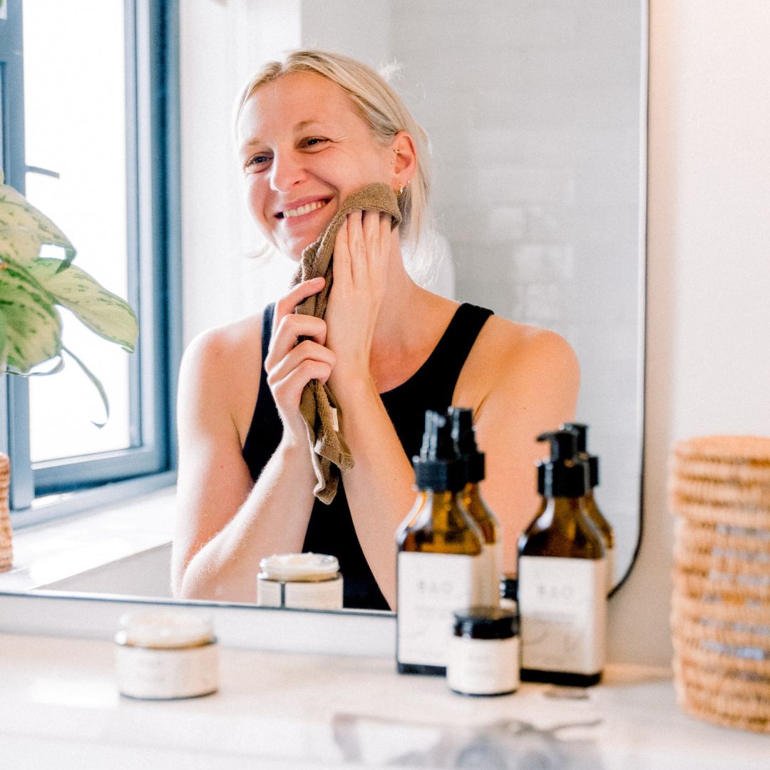 Woman smiling while wiping her face with a towel at a bathroom mirror with BAO products nearby