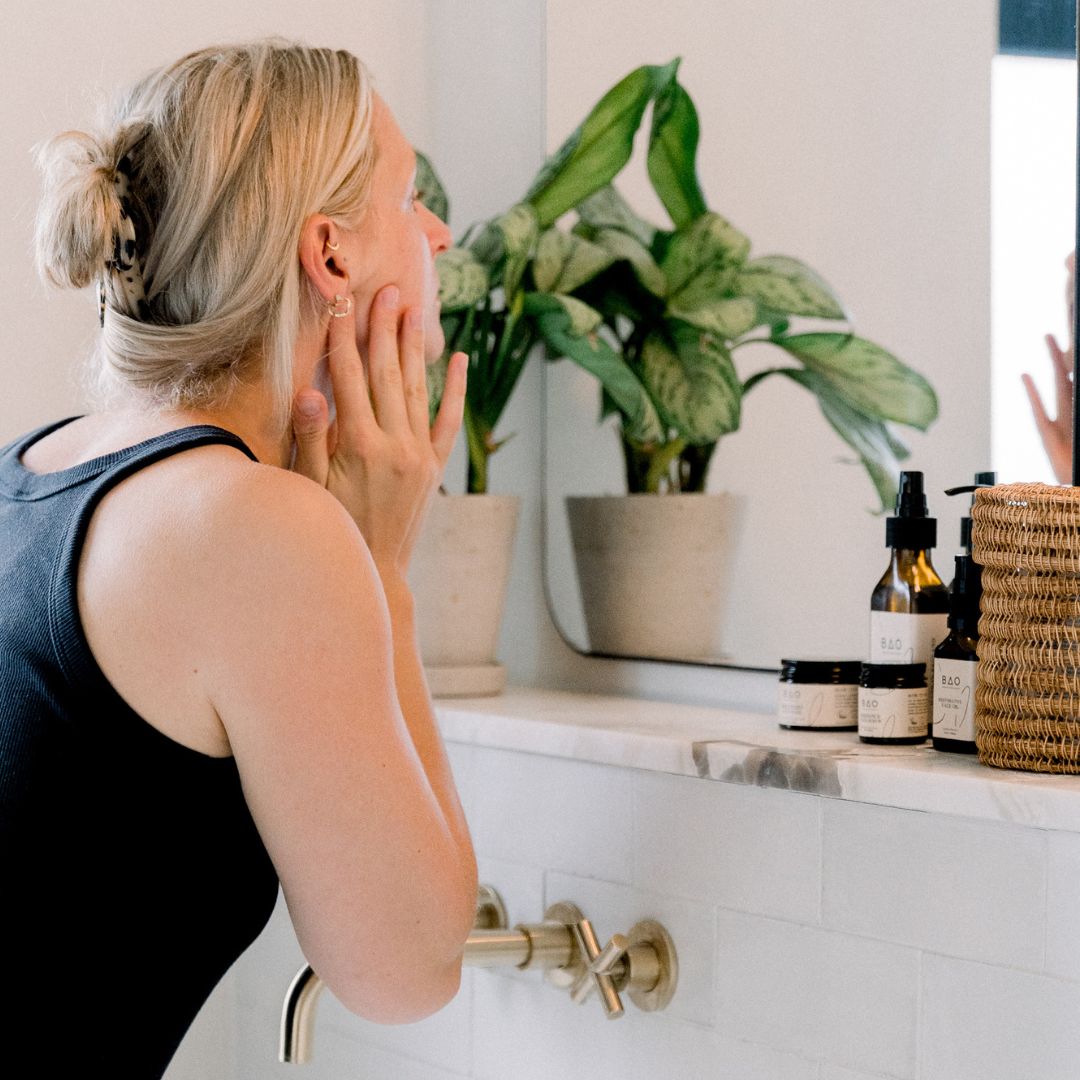 Woman applying BAO Restorative Face Oil to her face in front of a bathroom mirror