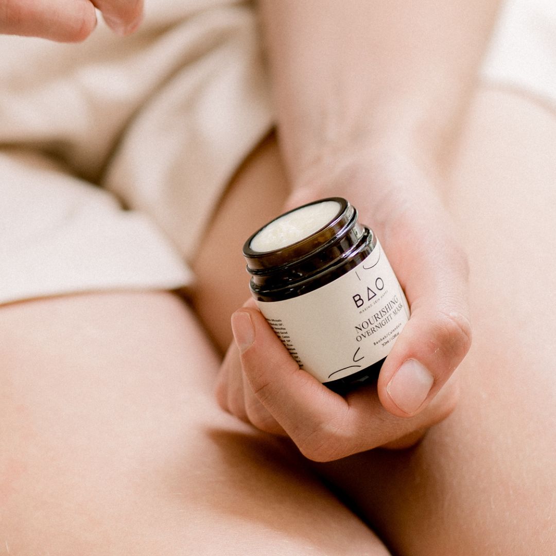 Hand holding an open jar of BAO Nourishing Overnight Mask in a bedroom setting