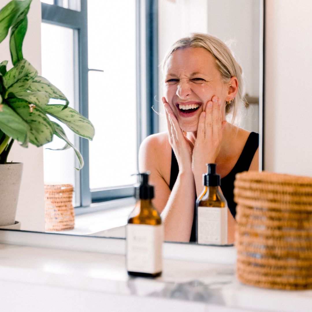 Woman laughing while touching her face at a bathroom mirror with BAO Refreshing Face Wash bottles nearby
