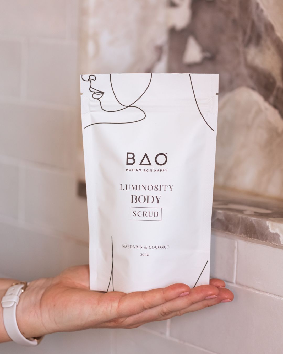 BAO Luminosity Body Scrub pouch held in an open palm against shower tiles