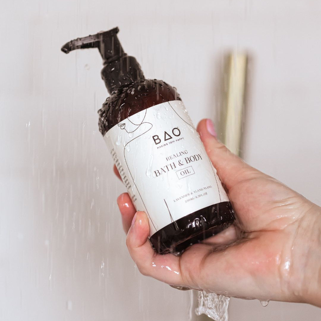 Hand holding BAO Healing Bath &amp; Body Oil bottle against a bathroom wall with incense sticks